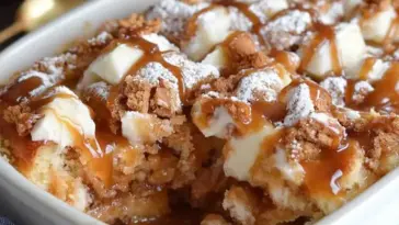Baked caramel apple cinnamon roll dump cake in a white dish, topped with powdered sugar, caramel drizzle, and soft cinnamon roll chunks.