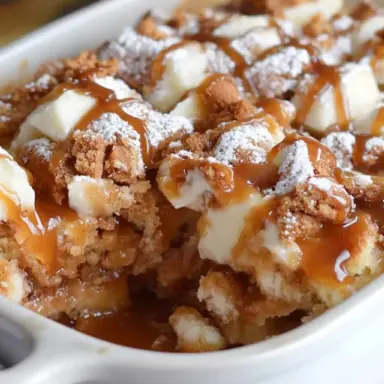 Close-up of a caramel apple cinnamon roll dump cake in a white baking dish, topped with gooey caramel drizzle, powdered sugar, and baked cinnamon roll pieces.
