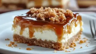 Close-up of a thick caramel cheesecake bar with creamy layers, a graham cracker base, rich caramel glaze, and brown sugar crumble on top, served on a white plate.