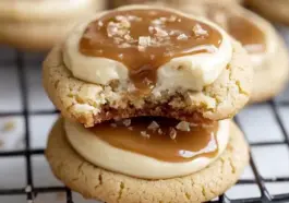 Stacked caramel cheesecake cookies on a cooling rack, topped with creamy caramel and flaky sea salt, showing a gooey cheesecake filling inside.