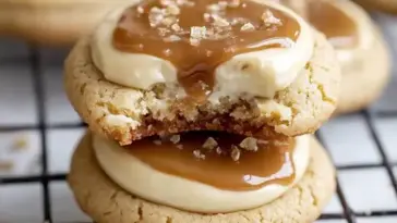 Stacked caramel cheesecake cookies on a cooling rack, topped with creamy caramel and flaky sea salt, showing a gooey cheesecake filling inside.