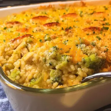 Close-up of cheesy chicken broccoli rice casserole in a white baking dish, with golden melted cheese, broccoli florets, and rice scooped onto a spoon.