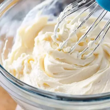 Close-up of creamy homemade cream cheese frosting in a glass bowl with a whisk.
