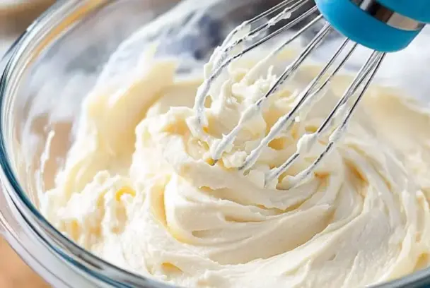 Whisk blending fluffy cream cheese frosting in a glass bowl—perfect for topping cakes and cupcakes.