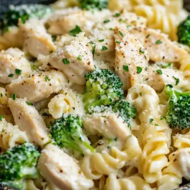 A close-up of creamy rotini pasta mixed with chunks of golden seared chicken and vibrant broccoli, coated in a rich, cheesy cream sauce and topped with fresh parsley.