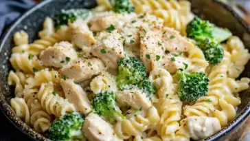 Top-down view of a bowl filled with creamy rotini pasta, seared chicken pieces, and broccoli florets, garnished with cracked pepper and chopped parsley.