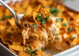 Cheesy Dorito Casserole being served from a white baking dish, showing melted cheese, crunchy nacho chips, and garnished with chopped parsley.