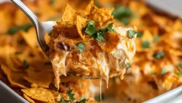 Cheesy Dorito Casserole being served from a white baking dish, showing melted cheese, crunchy nacho chips, and garnished with chopped parsley.