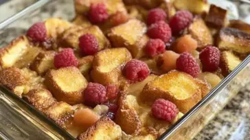 A glass baking dish filled with golden-brown baked French toast casserole, topped with fresh red raspberries and crispy toasted bread cubes on a granite countertop.