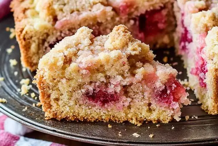 Slice of gluten free rhubarb crumb cake on a dark plate, showing golden crumble topping and vibrant baked rhubarb pieces.