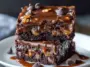 Two gooey chocolate caramel turtle cake bars stacked on a white plate, dripping with melted caramel and topped with chocolate chips and chopped pecans.