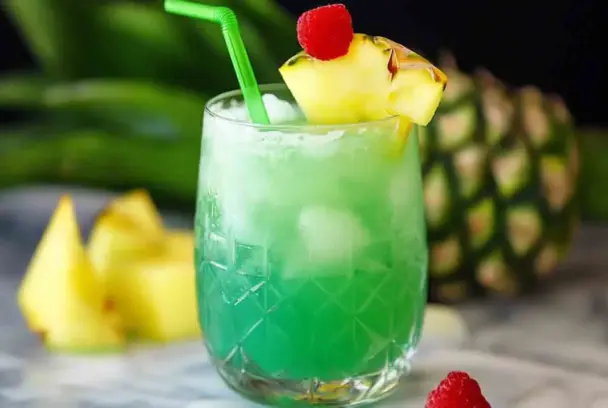 Bright green non-alcoholic tropical mocktail served in a patterned glass with ice, garnished with pineapple and a raspberry, with a green straw—ideal for Hawaiian-themed parties or summer drinks.