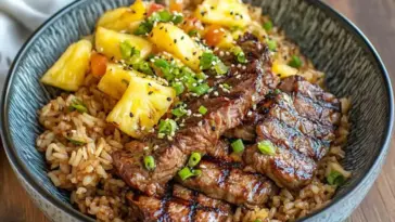 Close-up of a bowl filled with grilled teriyaki beef, golden pineapple chunks, and garlic fried rice, topped with sesame seeds and chopped green onions.