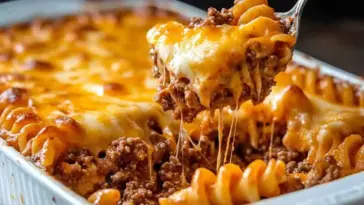 Close-up of a cheesy hamburger casserole being scooped from a baking dish, showing spiral pasta, seasoned ground beef, and melted golden cheese strands.