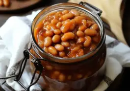 A glass jar filled with home canned baked beans in a thick, sweet molasses sauce, sitting on a kitchen towel with a wooden spoon in the background.