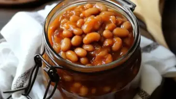 A glass jar filled with home canned baked beans in a thick, sweet molasses sauce, sitting on a kitchen towel with a wooden spoon in the background.