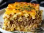 Close-up of a slice of Mac and Cheese Meatloaf Casserole on a white plate, showcasing creamy macaroni, seasoned beef, and golden melted cheese topping with herb garnish.