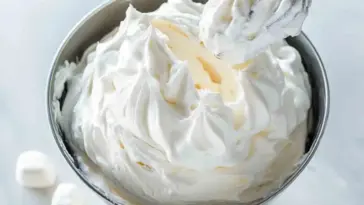 Wide view of whipped homemade marshmallow frosting in a stainless steel mixing bowl with peaks and a whisk, ready for decorating desserts