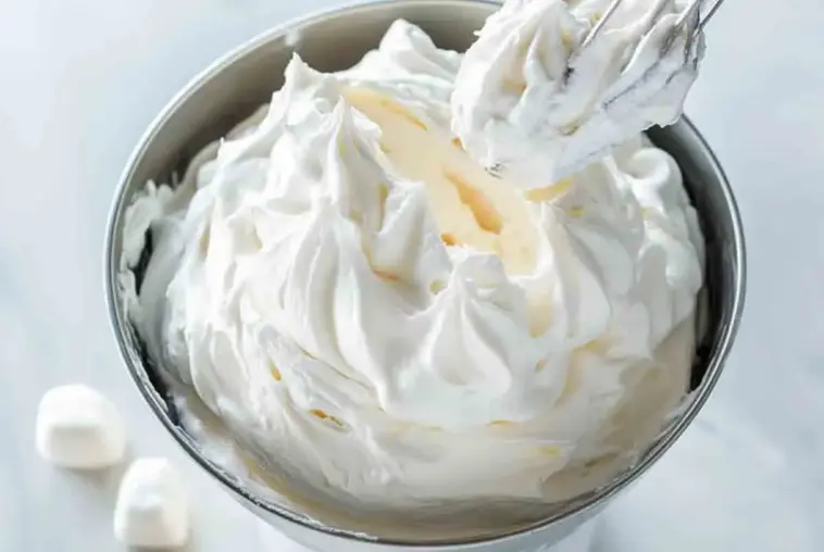 Wide view of whipped homemade marshmallow frosting in a stainless steel mixing bowl with peaks and a whisk, ready for decorating desserts