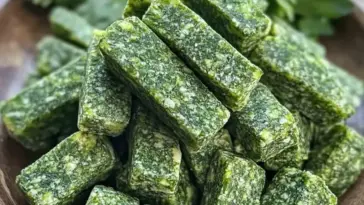Stack of homemade green parsley and mint dog treats in rectangular shapes, arranged on a wooden plate with fresh herbs in the background.