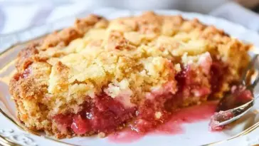 A slice of rhubarb dump cake with golden crumb topping and juicy pink rhubarb filling, served on an elegant white and gold-trimmed plate with a spoon.