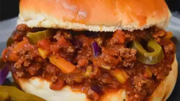 Close-up of a loaded Sloppy Joe sandwich on a soft bun, filled with seasoned ground beef, melted cheddar, diced onions, and sliced jalapeños.