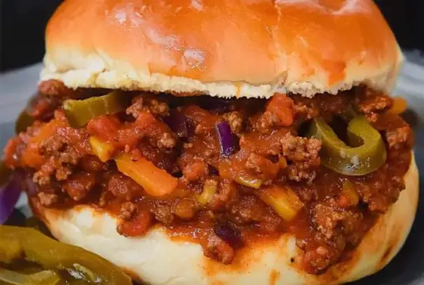 Close-up of a loaded Sloppy Joe sandwich on a soft bun, filled with seasoned ground beef, melted cheddar, diced onions, and sliced jalapeños.