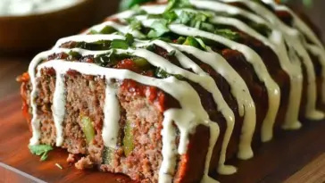 Close-up of sliced jalapeño popper meatloaf on a wooden board, filled with cheese and jalapeños, topped with ranch dressing and chopped fresh herbs.