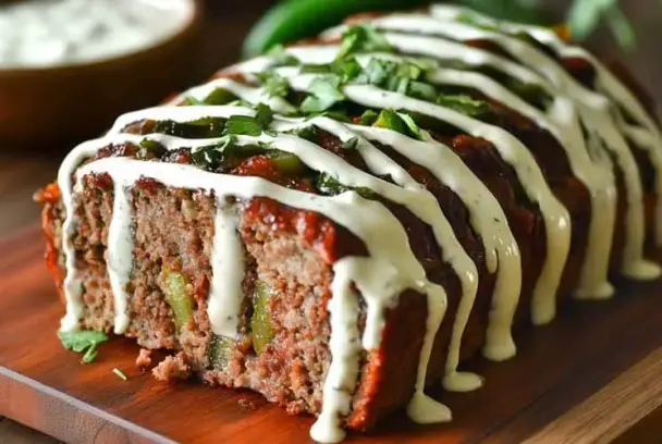Close-up of sliced jalapeño popper meatloaf on a wooden board, filled with cheese and jalapeños, topped with ranch dressing and chopped fresh herbs.
