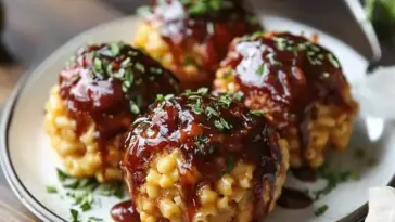 Close-up of four golden mac and cheese bombs filled with pulled pork and topped with maple bourbon BBQ sauce, garnished with fresh herbs on a white plate.