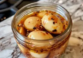 Close-up of spicy pickled eggs in a glass jar filled with chili flake and sesame seed brine, placed on a marbled kitchen counter.