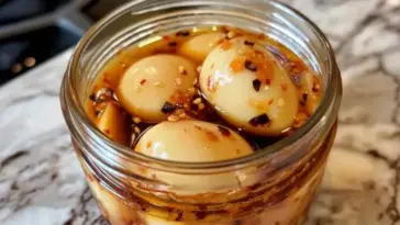 Close-up of spicy pickled eggs in a glass jar filled with chili flake and sesame seed brine, placed on a marbled kitchen counter.