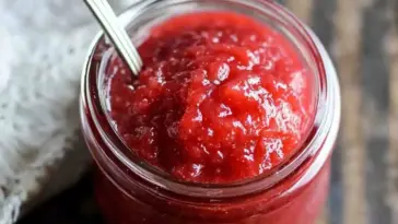 Close-up of a glass jar filled with thick, glossy homemade strawberry rhubarb sauce, with a spoon inside. The sauce is bright red and richly textured.