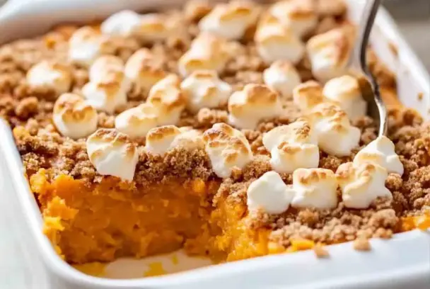 Close-up image of sweet potato casserole in a white baking dish, with a golden brown marshmallow topping and crisp pecan streusel, with a serving scooped out to reveal the creamy sweet potato base.