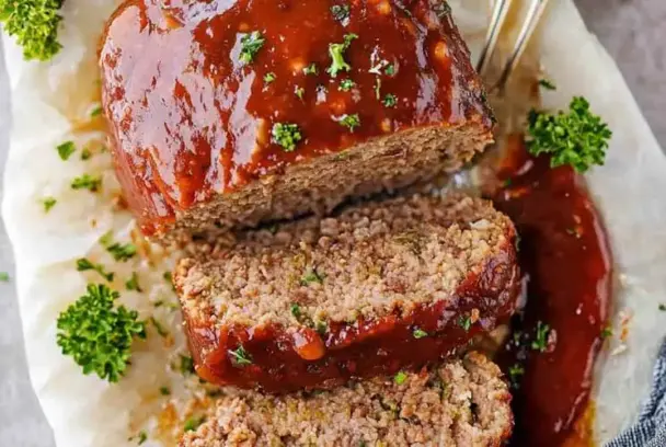 Overhead view of sliced meatloaf covered in a glossy ketchup glaze, garnished with parsley and served on parchment paper – a juicy homemade meatloaf.