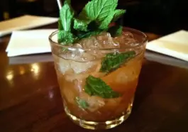 Glass filled with a classic mint julep cocktail, garnished with fresh mint sprigs and packed with crushed ice, served on a wooden table.