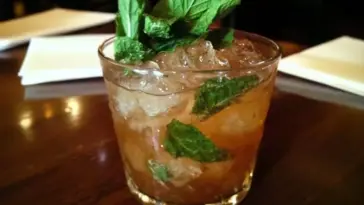 Glass filled with a classic mint julep cocktail, garnished with fresh mint sprigs and packed with crushed ice, served on a wooden table.