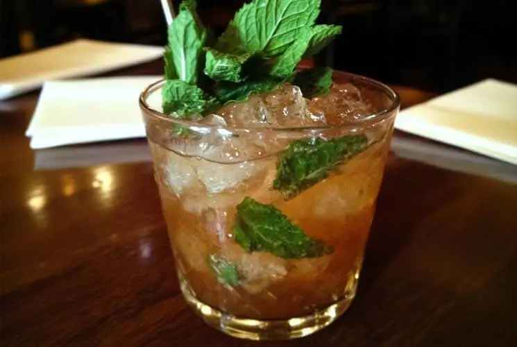 Glass filled with a classic mint julep cocktail, garnished with fresh mint sprigs and packed with crushed ice, served on a wooden table.