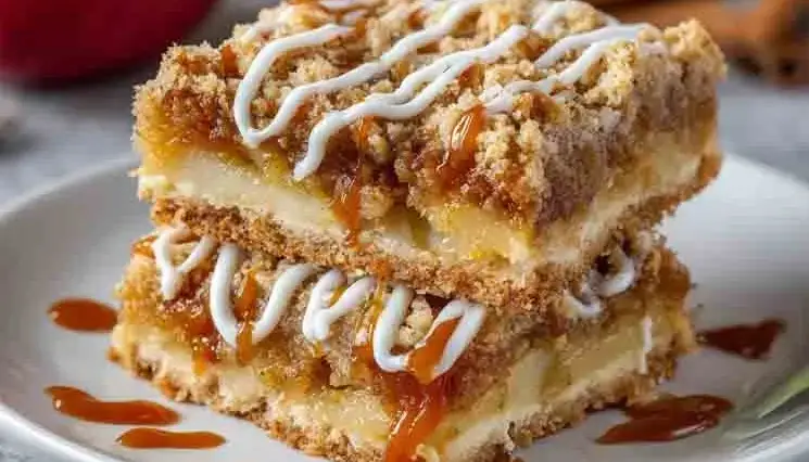 Stack of apple pie bars with caramel drizzle and icing on a white plate, showing layers of apple filling and golden crumble topping.
