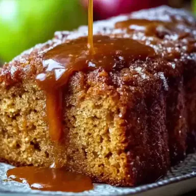 Close-up of a moist apple sauce cake slice topped with caramel glaze, served on a rustic plate with fresh apples in the background.