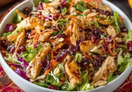 Overhead shot of Asian Chicken Cranberry Salad in a white bowl, featuring shredded chicken, dried cranberries, sliced almonds, carrots, purple cabbage, and sesame seeds on a colorful tablecloth.