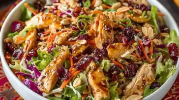 Overhead shot of Asian Chicken Cranberry Salad in a white bowl, featuring shredded chicken, dried cranberries, sliced almonds, carrots, purple cabbage, and sesame seeds on a colorful tablecloth.