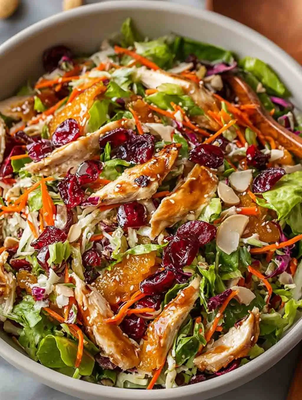 Close-up of a colorful Asian Chicken Cranberry Salad with shredded chicken, dried cranberries, mandarin oranges, sliced almonds, and shredded cabbage in a white bowl.