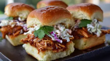 Close-up of BBQ chicken sliders topped with creamy coleslaw and fresh cilantro, served on golden toasted Hawaiian rolls on a dark platter.