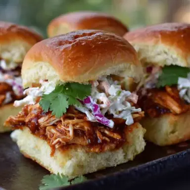 Close-up of BBQ chicken sliders topped with creamy coleslaw and fresh cilantro, served on golden toasted Hawaiian rolls on a dark platter.