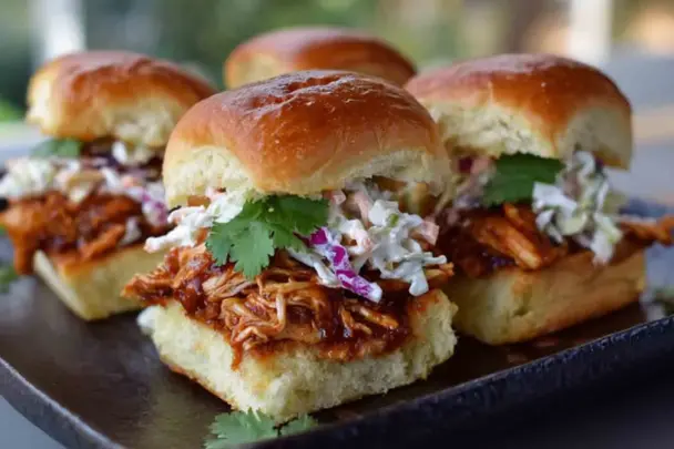 Close-up of BBQ chicken sliders topped with creamy coleslaw and fresh cilantro, served on golden toasted Hawaiian rolls on a dark platter.