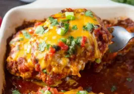 Close-up of a cheesy baked salsa chicken breast being lifted from a casserole dish, topped with melted cheddar cheese, fresh cilantro, diced red peppers, and green onions, surrounded by rich tomato salsa.