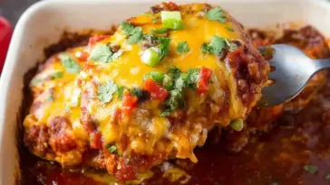 Close-up of a cheesy baked salsa chicken breast being lifted from a casserole dish, topped with melted cheddar cheese, fresh cilantro, diced red peppers, and green onions, surrounded by rich tomato salsa.
