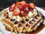 A close-up of two golden Belgian waffles topped with whipped cream, fresh strawberries, powdered sugar, and maple syrup on a white plate with a fork.