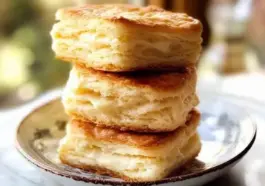 Three stacked golden brown flaky biscuits on a ceramic plate, with visible buttery layers and a soft, fluffy texture.